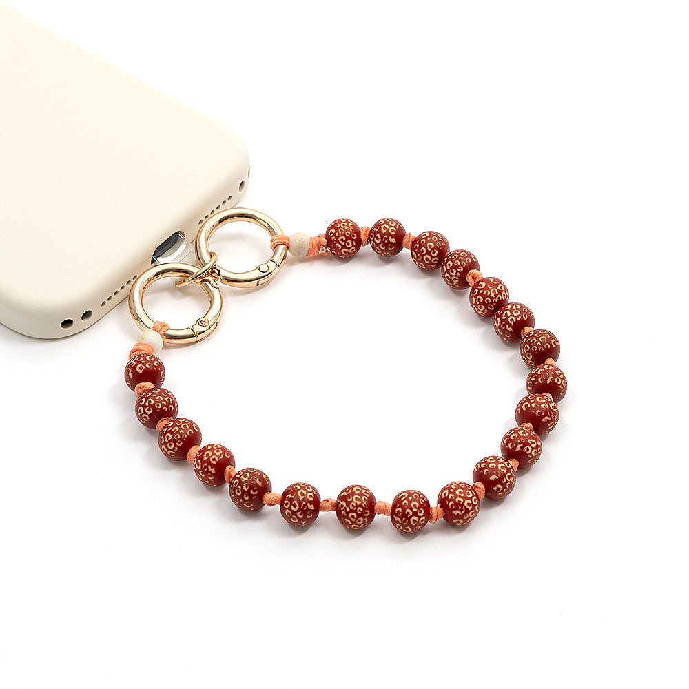 Roter Handy-Anhänger aus Perlen mit goldenen Ringen und handgefertigten Holzperlen von Alexandra Renke, befestigt an einer weißen Handyhülle im Leo-Muster-Stil, vor weißem Hintergrund.