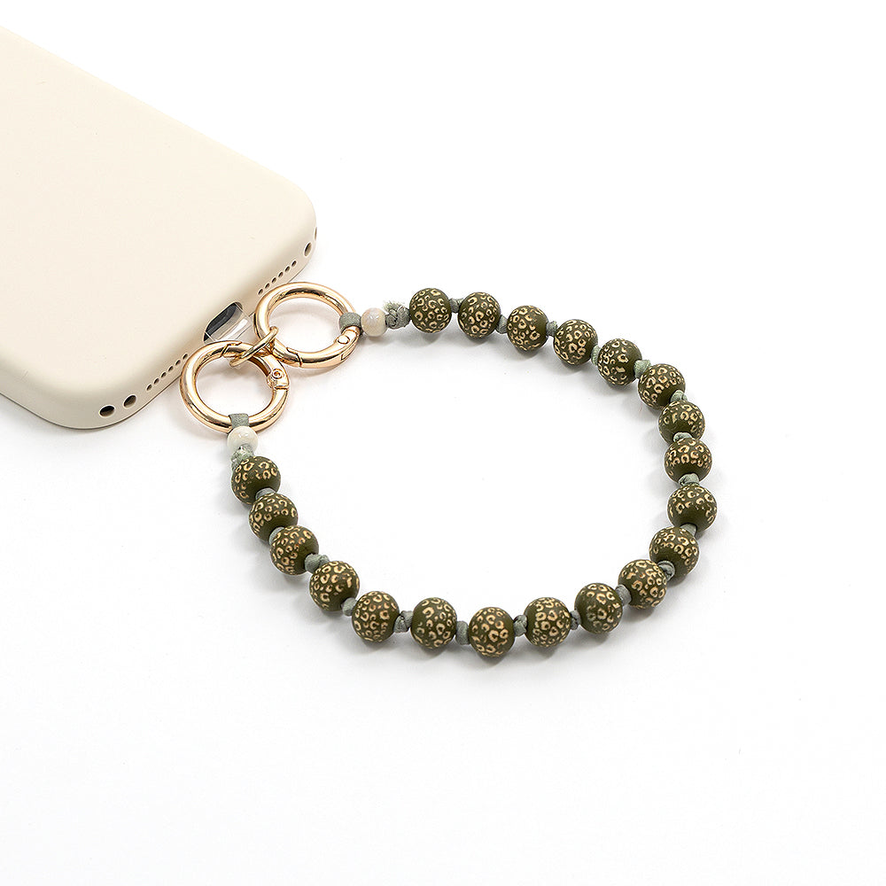 Perlenbesetztes Handyband mit goldenen Akzenten und Holzperlen, mit dezentem Löwenmuster, handgefertigt von Alexandra Renke und an einer beigen Handyhülle auf weißem Hintergrund befestigt.