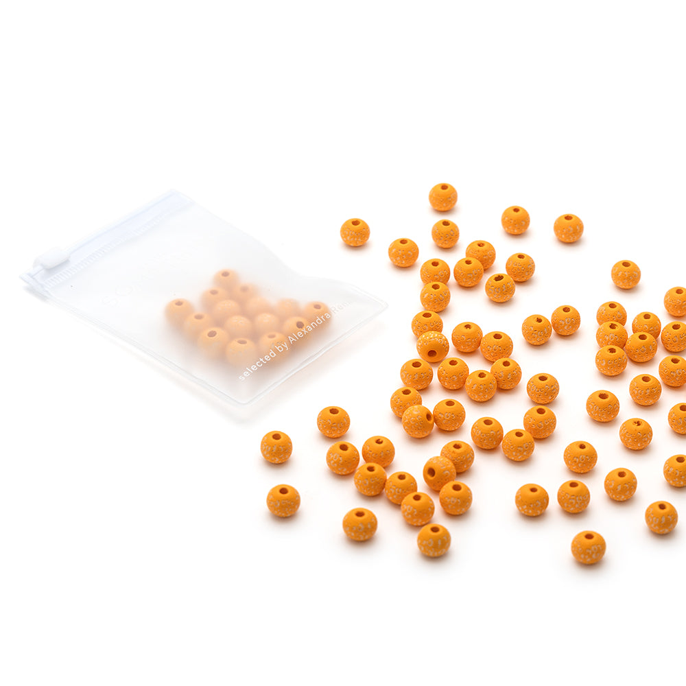 Kleine orangefarbene Holzperlen von Alexandra Renke verstreut neben einer transparenten Reißverschlusstasche auf einer weißen Fläche - ideal für Leo-Muster und handgefertigte Projekte.