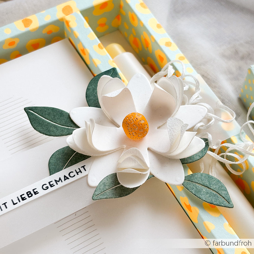 Eine weiße Papierblume mit grünen Blättern und einer gelben Mitte ruht auf einer Geschenkbox mit einem Blumenmuster im Leo-Stil, die mit handgefertigten Holzperlen verziert ist und so einen eleganten Akzent setzt.