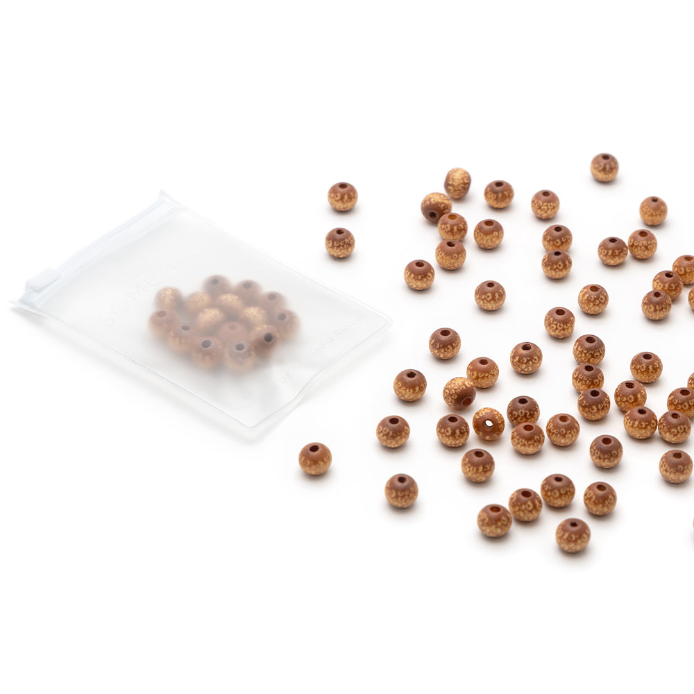 Kleine runde braune Holzperlen von Alexandra Renke, einige verstreut und einige in einer klaren Plastiktüte, ausgestellt auf weißem Hintergrund.