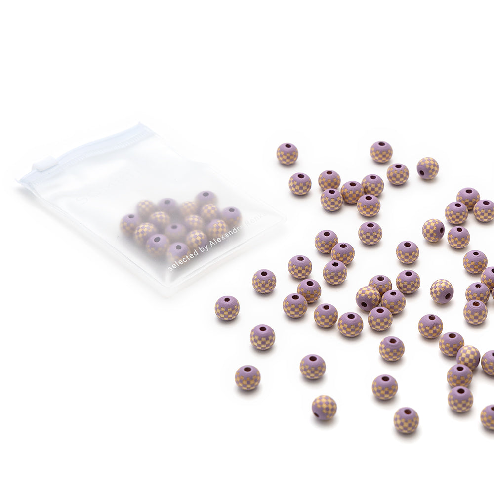Eine durchsichtige Tasche und verstreute Alexandra Renke handgefertigte Holzperlen mit einem violetten Boden, goldenen gepunkteten grafischen Mustern, und einem großen Öhr auf weißem Hintergrund.