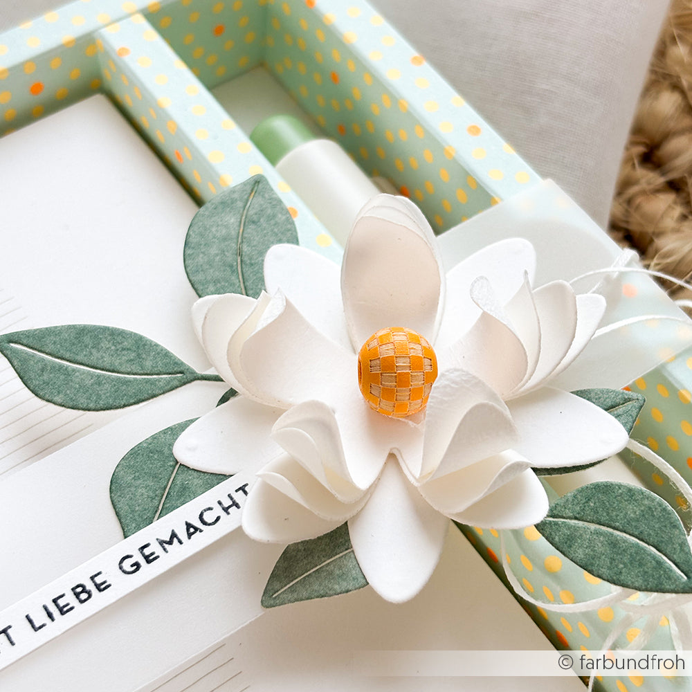 Auf einer weiß-grünen Schachtel befindet sich eine handgefertigte Blume aus weißem Papier mit grünen Blättern und einer orangefarbenen Mitte, die durch Holzperlen akzentuiert wird.