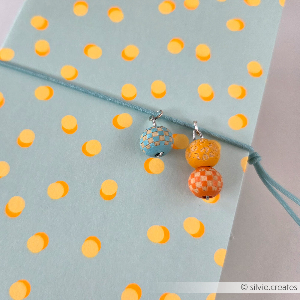 Drei gemusterte Holzperlen in Blau und Orange liegen auf einem hellblauen Notizbuch mit gelben Punkten und einem blauen Gummiband – ideal für Ihre Projekte mit handgefertigtem Bastelzubehör.