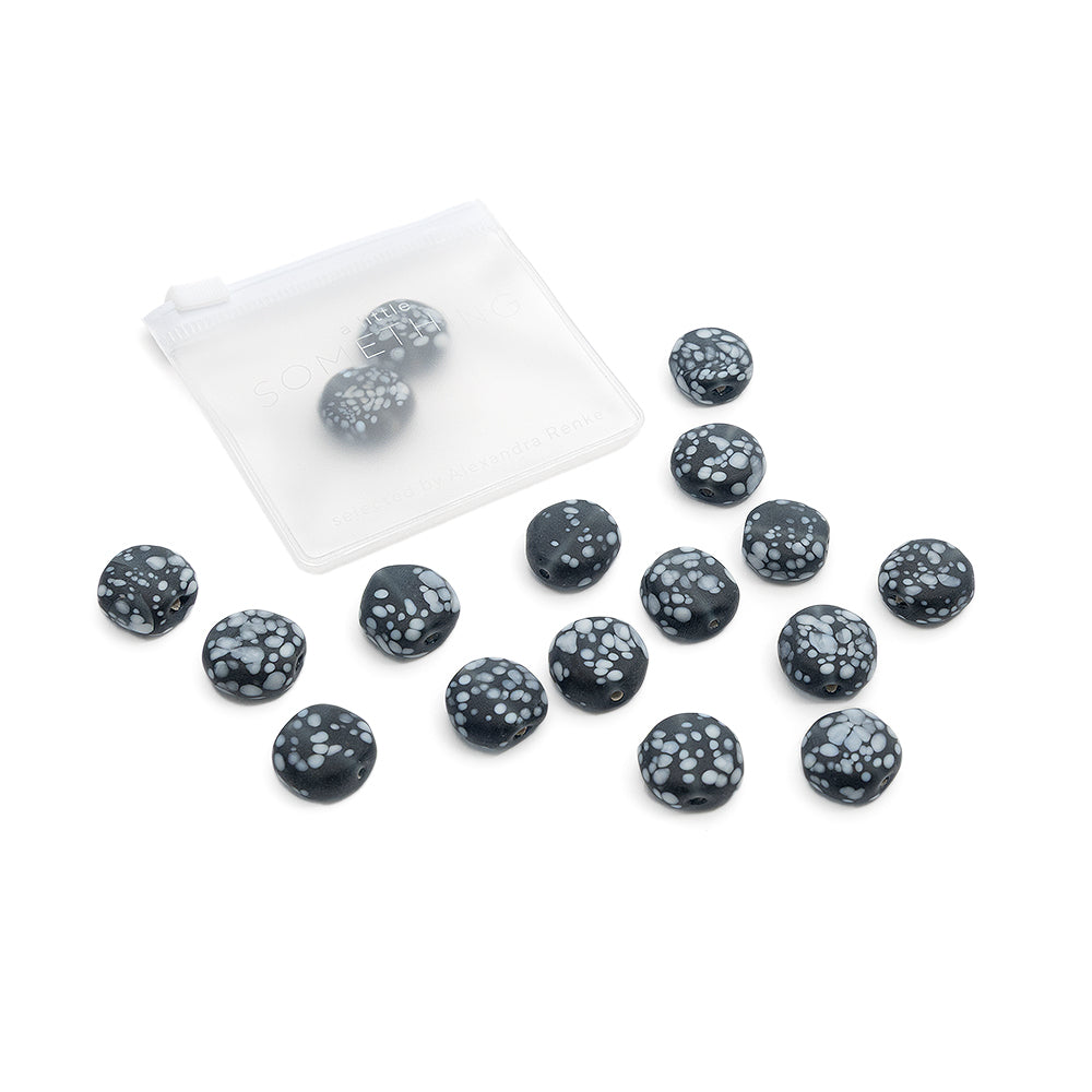 Vierzehn schwarze Steine mit weißen Sprenkeln liegen neben einem kleinen, durchscheinenden Säckchen, zusammen mit einem Set runder Glasperlen von Alexandra Renke für Ihr nächstes DIY-Schmuckprojekt.