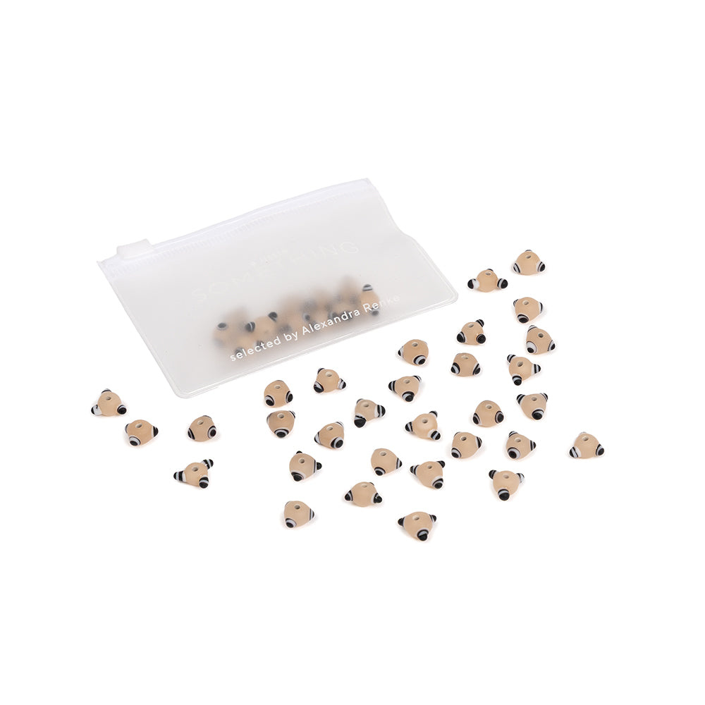 Kleine, beige, fischförmige Perlen – ideal für Handarbeiten und DIY-Projekte – liegen auf weißem Hintergrund neben einem durchscheinenden Beutel mit Reißverschluss verstreut.