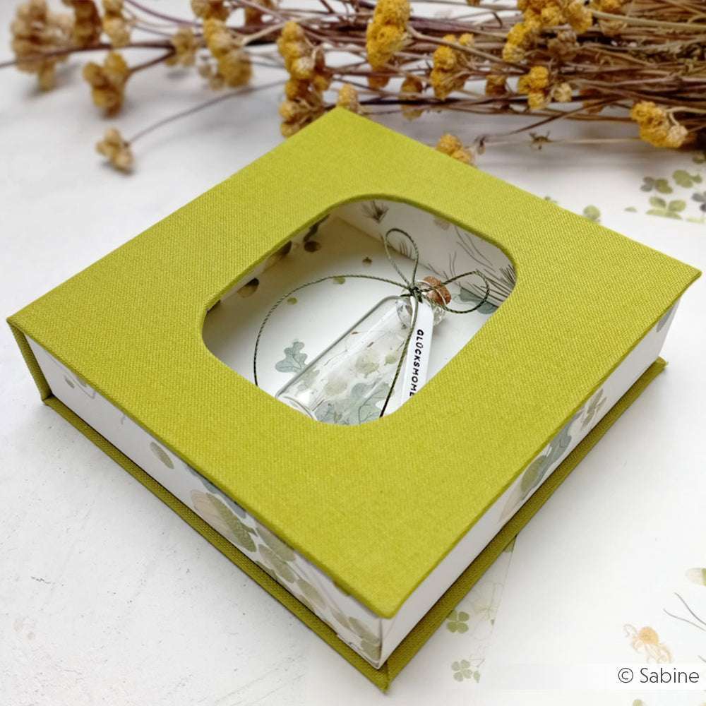 Die grüne PAPPcut-Geschenkbox von Alexandra Renke hat ein durchsichtiges Fenster, in dem ein kleines Glasfläschchen und Blumenschmuck zu sehen sind.