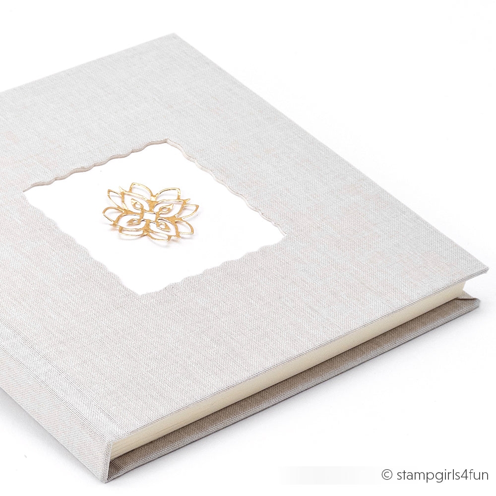 Ein hellgraues Hardcover-Notizbuch von Alexandra Renke, das mit Buchbinden und PAPPcut-Techniken hergestellt wurde, weist ein goldenes Blumenmuster auf dem Einband auf und wird elegant auf einer weißen Oberfläche präsentiert.