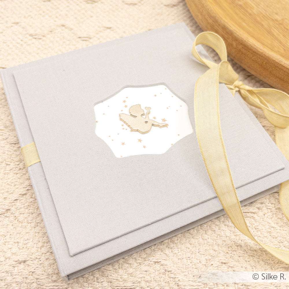 Hellgraues Fotoalbum von Alexandra Renke mit Meerjungfrauenmotiv und goldenem Band auf dem Einband, in Buchbindetechnik gefertigt, auf beiger Oberfläche.