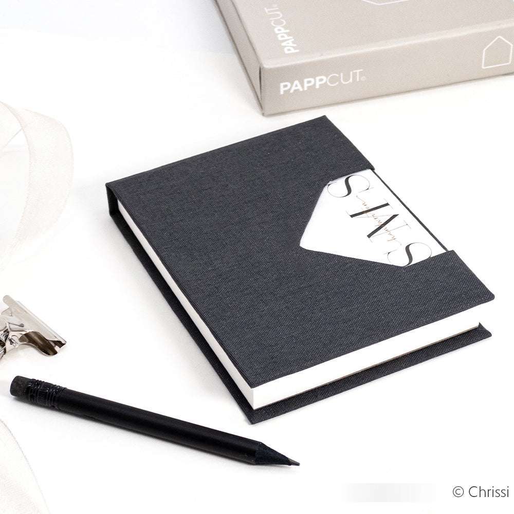 Schwarzes Hardcover-Notizbuch von Alexandra Renke mit einem Stift und einer Karte, die zum Teil in die Vordertasche gesteckt wird. Die hochwertige Buchbinden-Handarbeit wird auf einer weißen Oberfläche hervorgehoben.