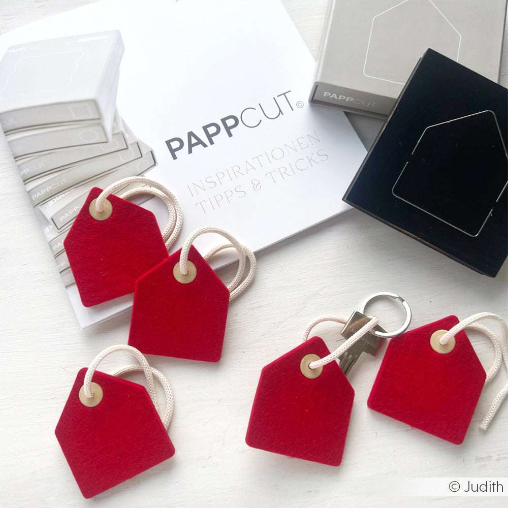 Rote Filzhaus-Schlüsselanhänger auf einem weißen Tisch mit Alexandra Renke-Verpackungen und einem Buchbinden-Heft im Hintergrund.
