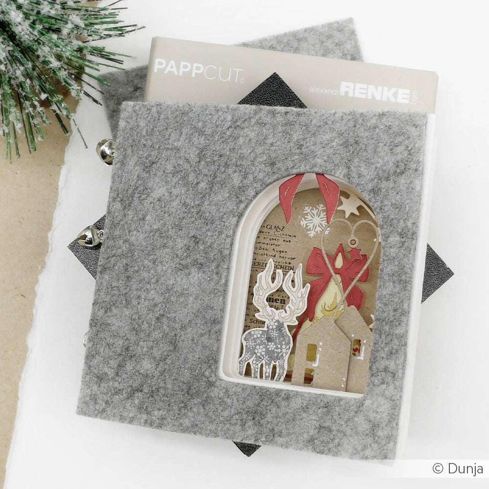Die graue Filz-Weihnachtskarte von Alexandra Renke, die mit den Techniken PAPPcut und Buchbinden hergestellt wurde, hat ein ausgeschnittenes Fenster, das ein Rentier, ein Haus und festliche Dekorationen im Inneren zeigt.