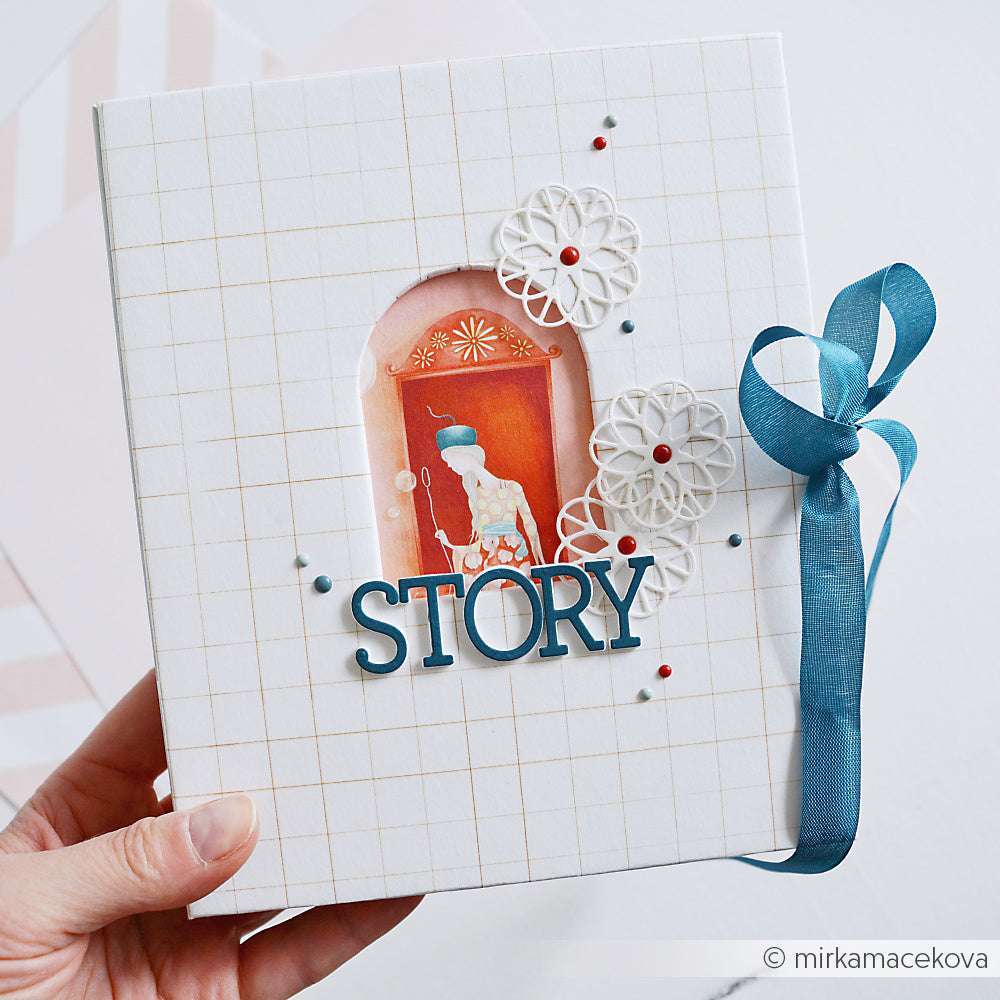 Eine Hand hält ein Sammelalbum von Alexandra Renke mit der Aufschrift "STORY" auf dem Einband, einem blauen Band an der Seite und verschlungenen Scherenschnittmustern.