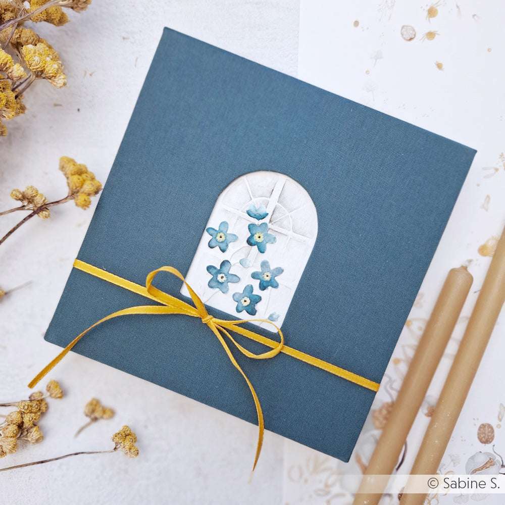 Alexandra Renke quadratische blaue Geschenkschachtel mit gelber Schleife und Fenstermotiv, hergestellt in Buchbindetechnik, ausgestellt zwischen Trockenblumen und Kerzen.