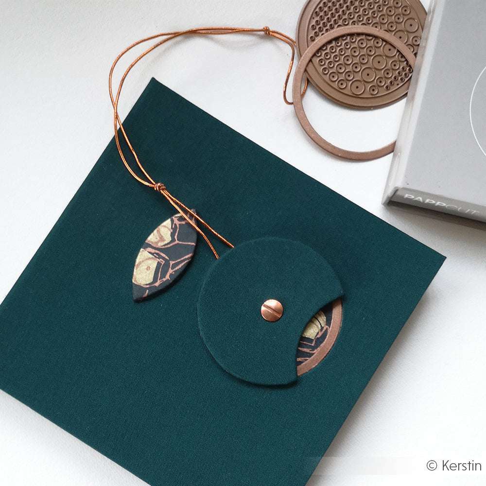 Flachdruck eines grünen Umschlags mit Knopfverschluss, Zierkordel und blattförmigem Anhänger, mit Akzenten, die mit den Techniken Alexandra Renke Buchbinden Stanze und PAPPcut Kreis hergestellt wurden.