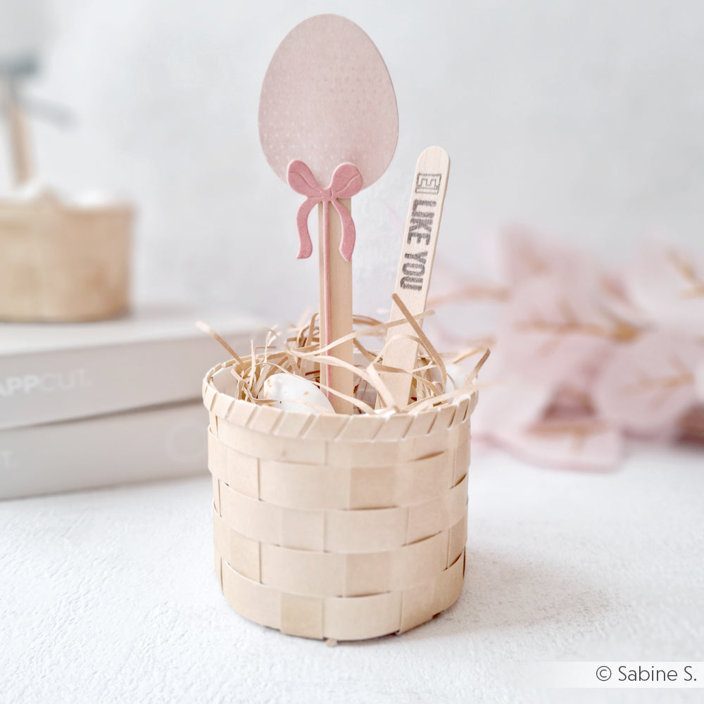 Kleines geflochtenes Körbchen mit rosa Eierdekoration und einem Alexandra Renke PAPPcuties Holzstab mit der Aufschrift "I LIKE YOU", eingebettet in eine Strohfüllung.