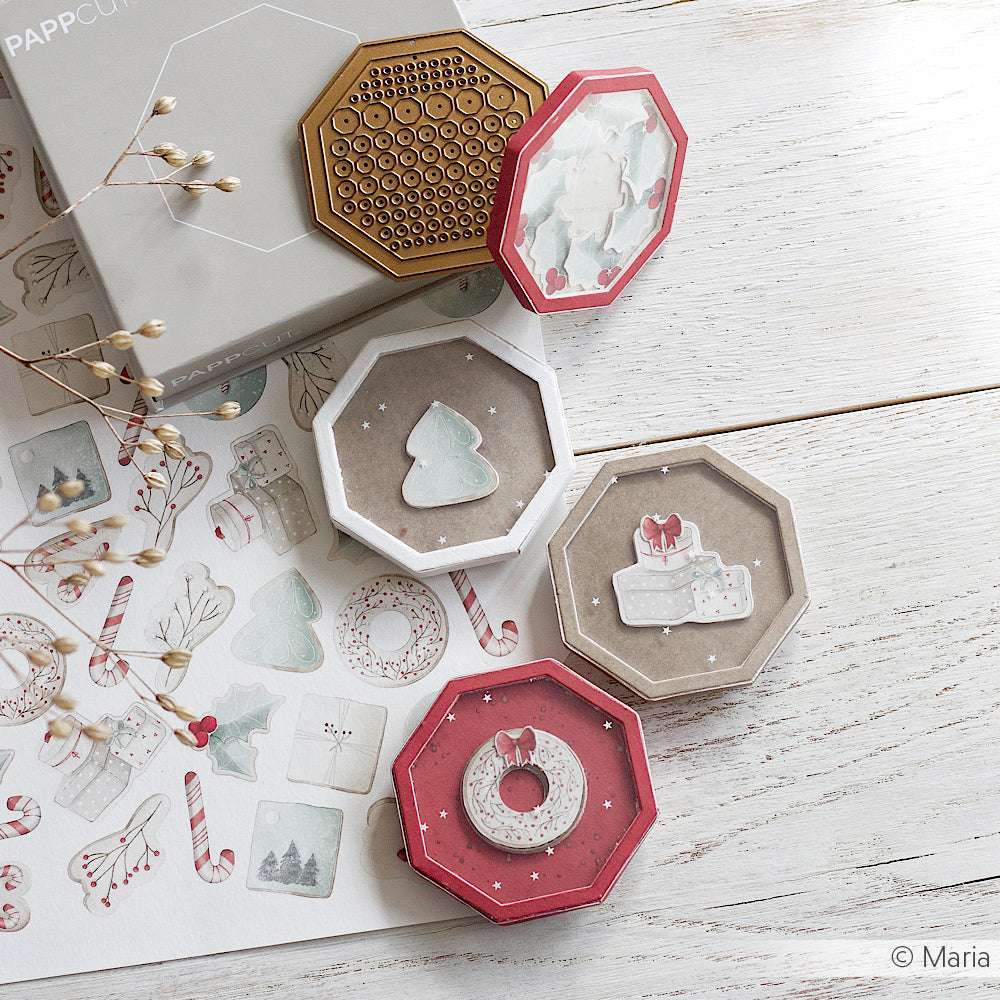 Vier achteckige weihnachtliche Geschenkboxen von Alexandra Renke mit festlichen Motiven und PAPPcut-Kanten stehen mit Aufklebern und einer grauen Schachtel auf einem weißen Holztisch - ideal zum Buchbinden oder kreativen Verpacken von Geschenken.