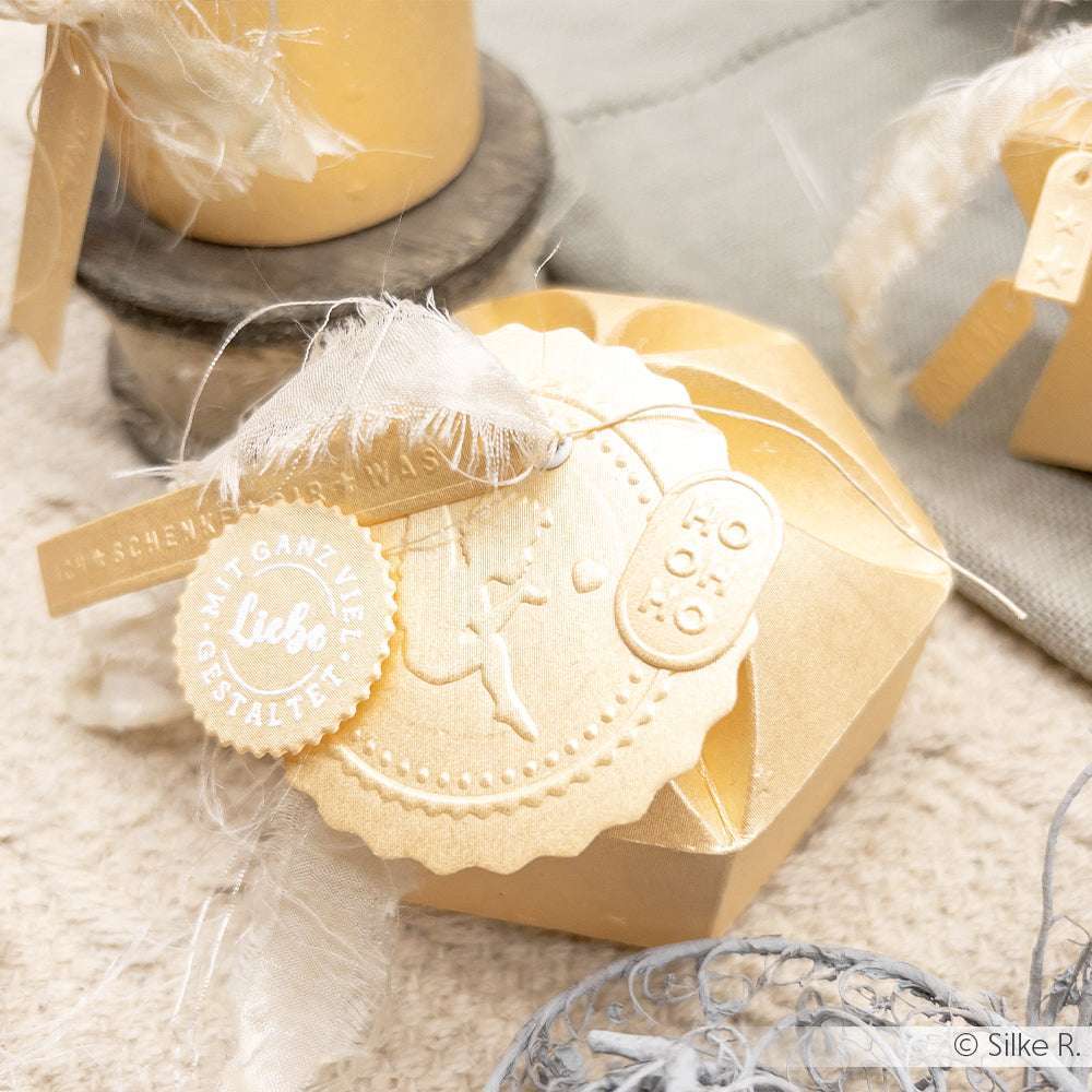 Goldfarbene Geschenkschachtel mit geprägtem Siegel, weißem Band und dekorativen Anhängern auf Schimmer-Effekt Papier (FSC-zertifiziert, 310 g/m²), dargestellt auf einer leicht strukturierten Oberfläche.