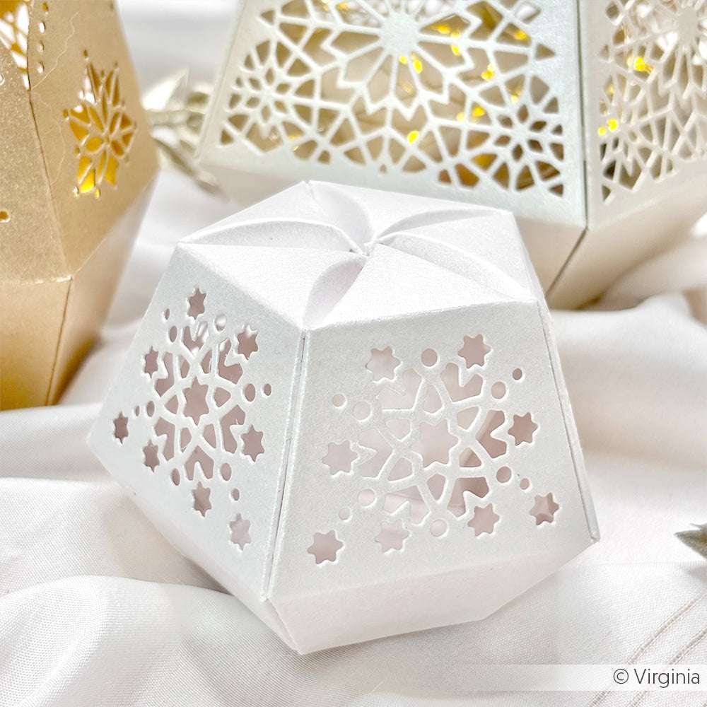 Weiße geometrische Papierlaterne aus hochwertigem Papier mit Sternen- und Schneeflockenausschnitten auf einer weißen Stoffoberfläche.