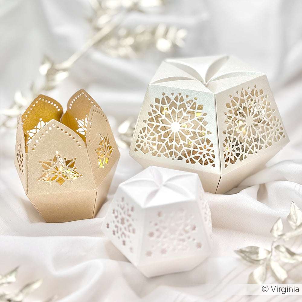 Drei dekorative, lasergeschnittene Papierlaternen in Weiß und Gold von ☆, gefertigt aus schimmerndem Papier, ruhen auf einem weichen weißen Stoffhintergrund - eine eindrucksvolle Präsentation von Papierkunst.