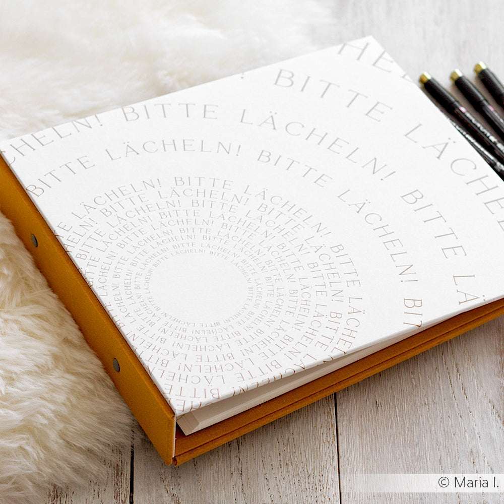 Ein weißer Alexandra-Renke-Ordner mit der Spiralaufschrift "BITTE LÄCHELN!", der sich perfekt für die Erstellung eines Tagebuchs eignet, liegt auf einer Holzunterlage, neben der Bleistifte liegen.