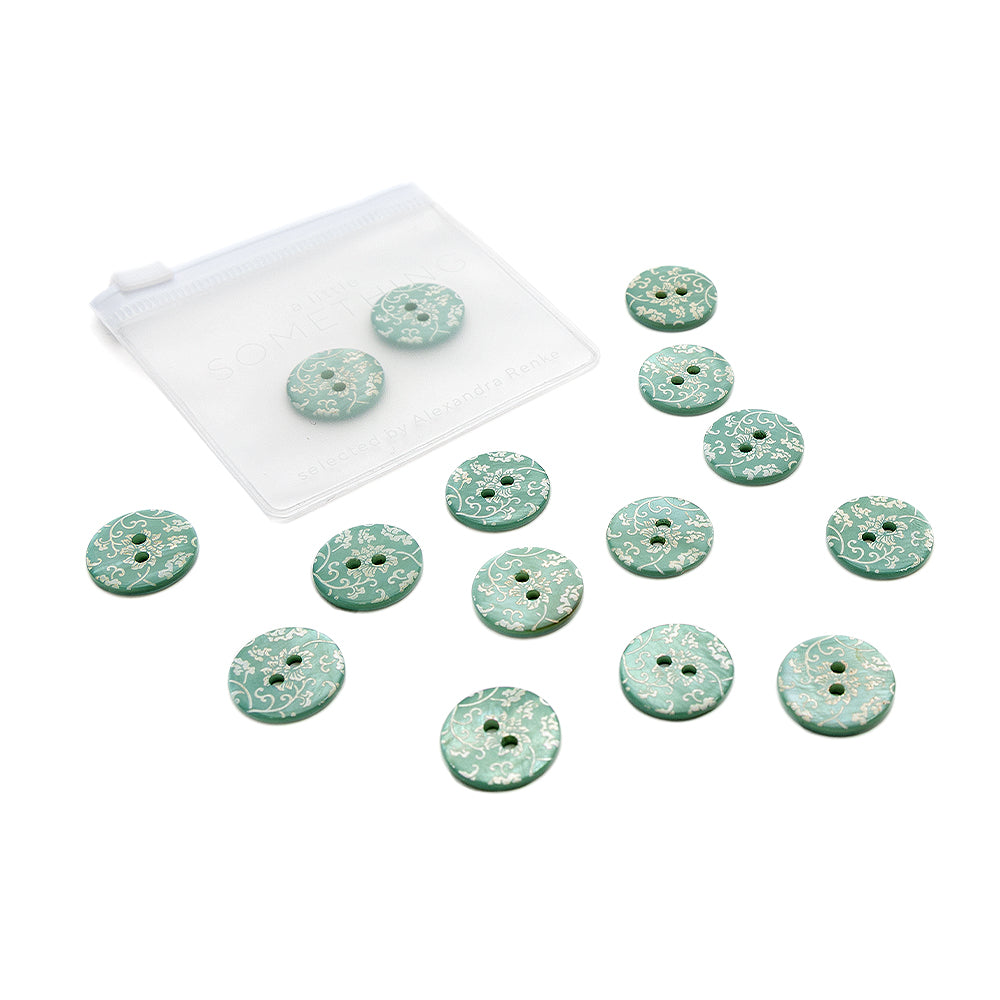 Ein Set aus 13 grünen, gemusterten Knöpfen von Alexandra Renke – einige davon mit zarten Blumenmotiven verziert – lag verstreut neben einem kleinen, durchsichtigen Beutel mit Reißverschluss.