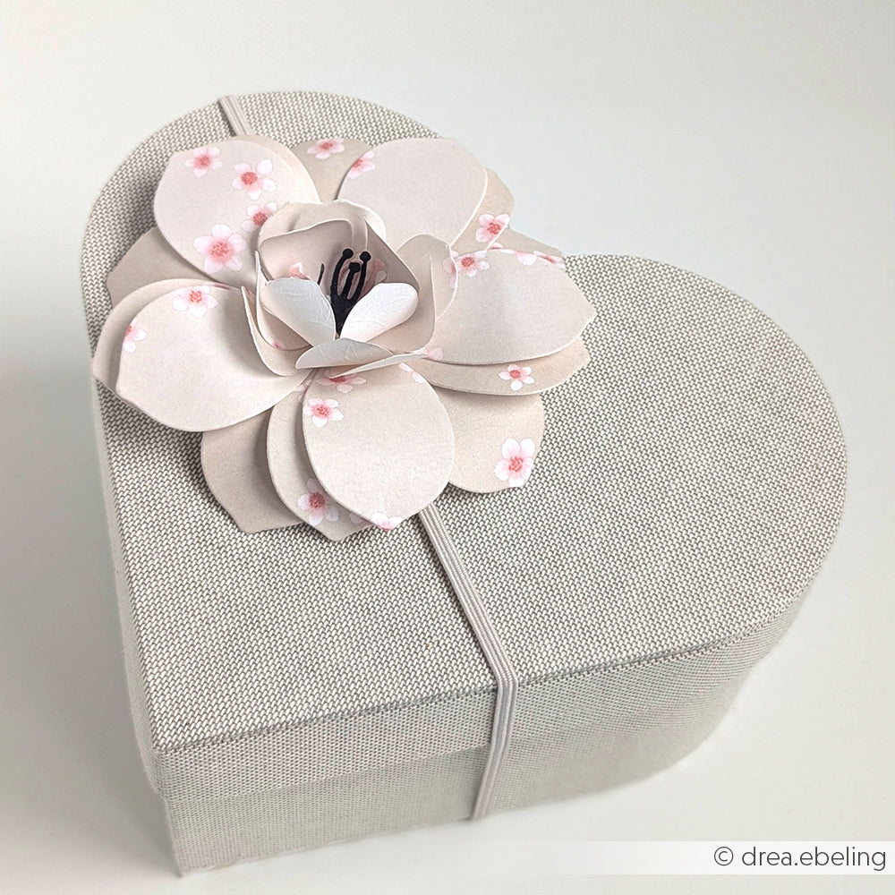 Herzförmige Geschenkbox mit einer großen, zarten Papierblume auf dem Deckel, hergestellt mit dem Alexandra Renke PAPPKit Herzbox Bastelset, vor einem schlichten weißen Hintergrund.