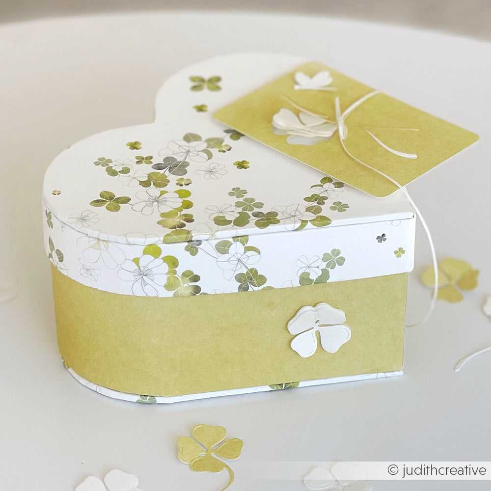 Herzförmige Geschenkverpackung mit grünem Kleemotiv und passendem Anhänger, hergestellt mit dem Alexandra Renke Herzbox Bastelset Geschenkverpackung DIY PAPPKit, ausgestellt auf einer weißen Fläche.