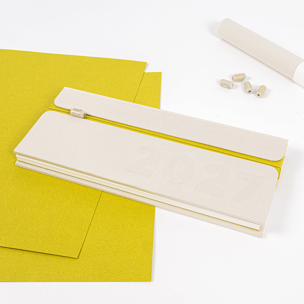 Rechteckiger Planer „2027“ mit beigem Einband auf gelbem Papier, Bleistift und kleinen runden Radiergummis daneben – perfekt, um dein Jahr zu organisieren oder mit dem vielseitigen PAPPKit deinen eigenen DIY-Kalender zu gestalten.