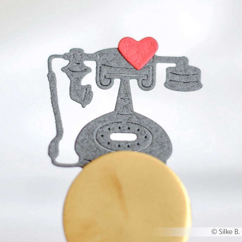 Plätzchen in Form eines grauen Vintage-Telefons, hergestellt mit dem Alexandra Renke Motivstanze Telefon Set, mit einem roten Herz auf der Spitze und vor einem hellen Hintergrund.