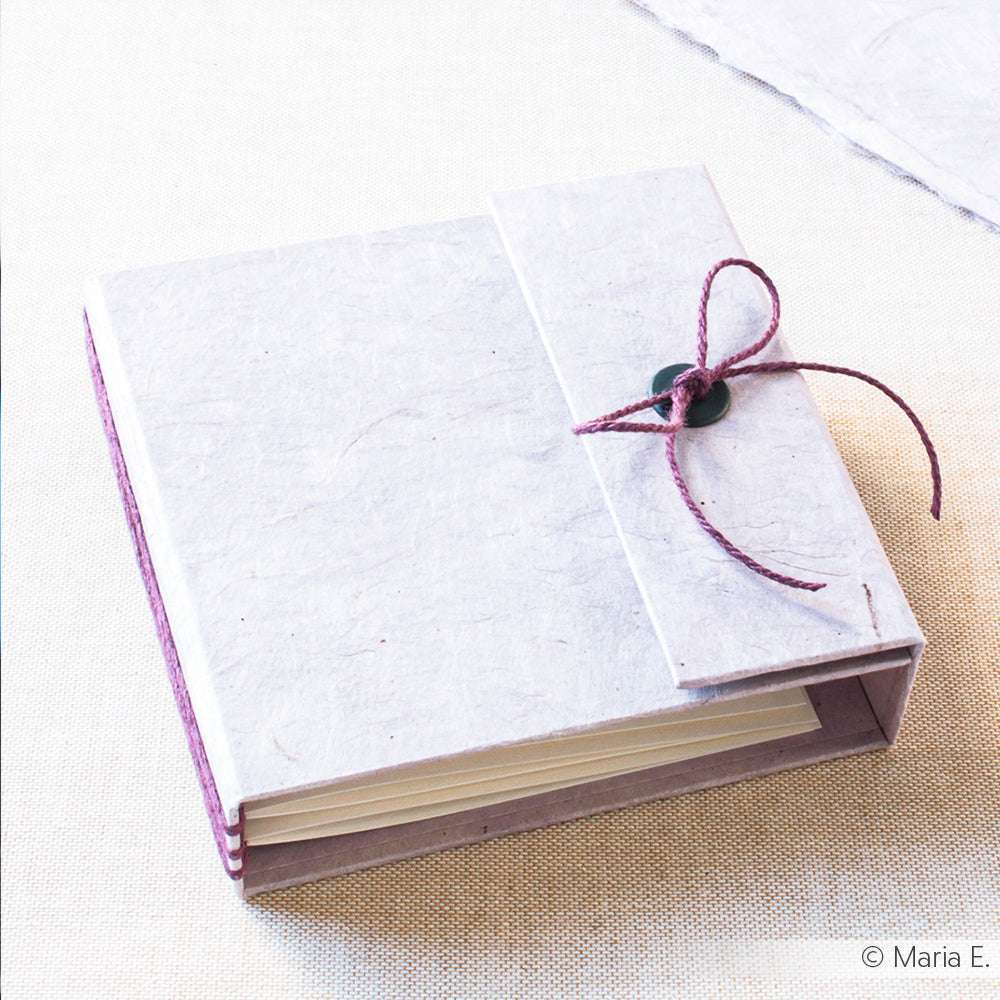 Quadratisches, handgefertigtes Buch von ☆ mit einer lila Schnur und einem Knopfverschluss, hergestellt mit Nepalpapier, auf einer leicht strukturierten Oberfläche.