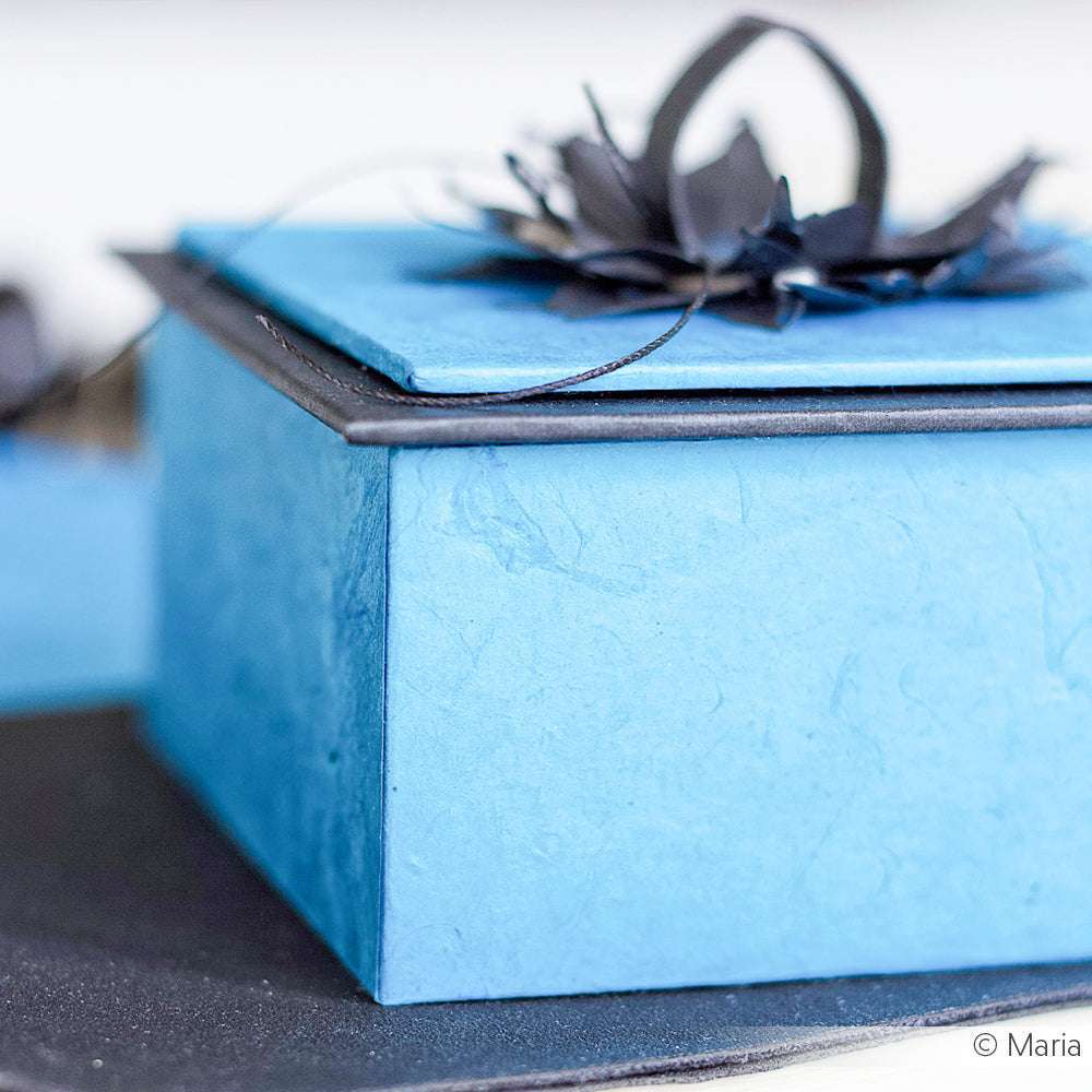 Eine ☆ blaue Geschenkbox mit einer dekorativen schwarzen Blume auf dem Deckel, hergestellt aus handgeschöpftem Nepalpapier, ausgestellt auf einer dunklen Oberfläche.