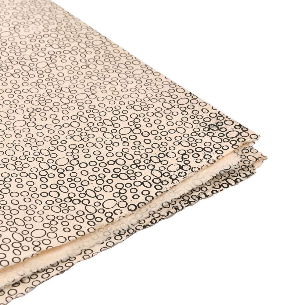 ☆ Nepalpapier in Beige mit unregelmäßigen schwarzen Kreisen, überlappen sich leicht. Das Papier ist an der Ecke gefaltet und liegt auf weißem Hintergrund - perfekt zum Geschenke Verpacken.