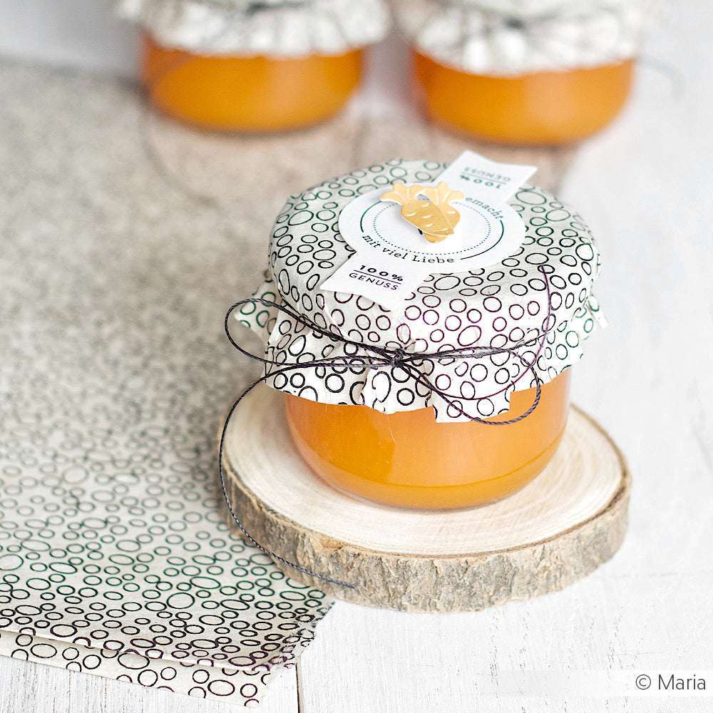 Ein Glas mit Orangenmarmelade von ☆ mit gemustertem Stoff und Nepalpapier steht auf einem Holzuntersetzer - ideal für Geschenke Verpacken. Zwei weitere ☆-Gläser erscheinen im Hintergrund.