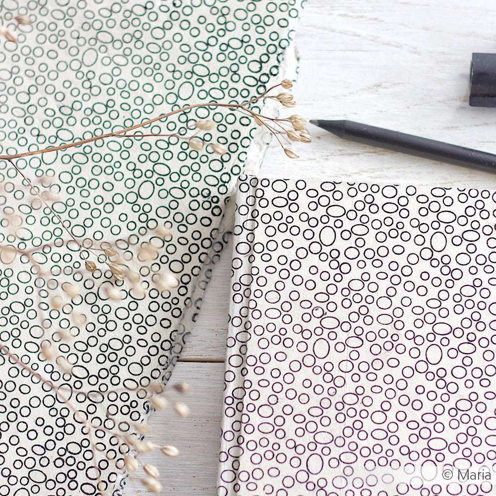 Zwei ☆ Nepalpapier-Notizbücher mit schwarzen ovalen Mustern, einem Stift und getrocknetem Gras auf einer weißen Holzoberfläche - ideal für Buchbinden-Projekte oder einzigartige Geschenke Verpacken-Inspirationen.