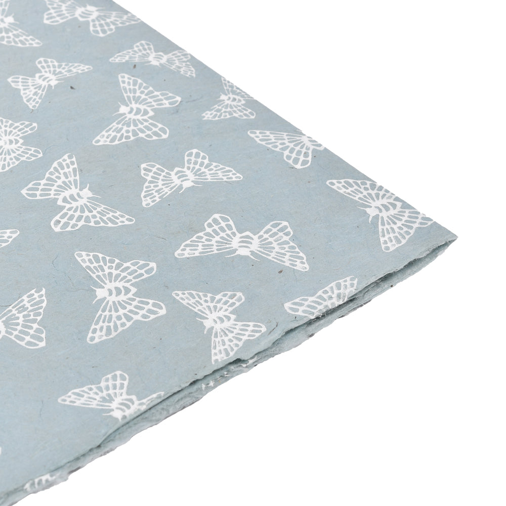 Hellblaues Nepalpapier mit einem sich wiederholenden weißen Schmetterlingsmuster, ideal zum Einpacken von Geschenken oder zum Buchbinden, abgebildet in einem Winkel vor weißem Hintergrund.