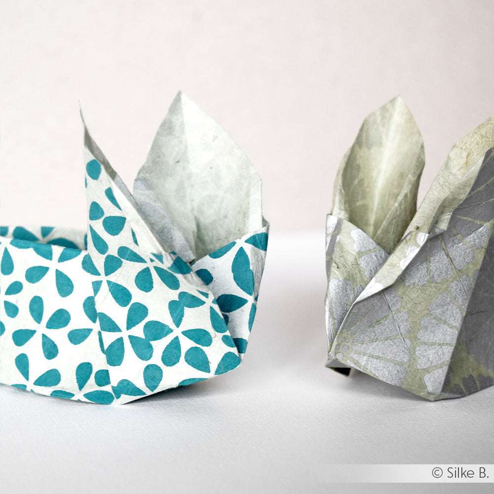 Zwei Origami-Hasen von ☆-einer mit blaugrünen Mustern auf Geschenkpapier, der andere grau auf Nepalpapier- sind auf einer weißen Fläche vor einem hellen Hintergrund angeordnet.