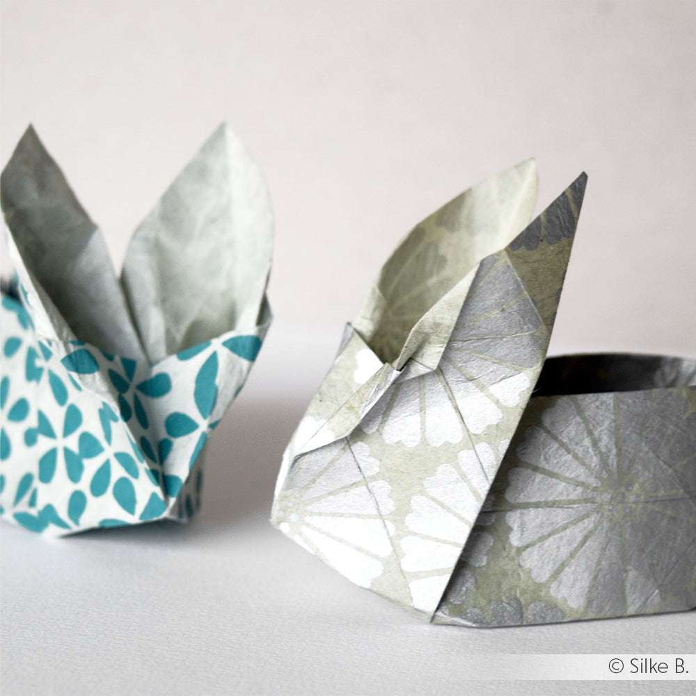 Zwei gemusterte Origamihasen von ☆, ein blauer und ein grauer, gefaltet aus Nepalpapier, sitzen auf einer weißen Fläche vor einem einfarbigen Hintergrund.