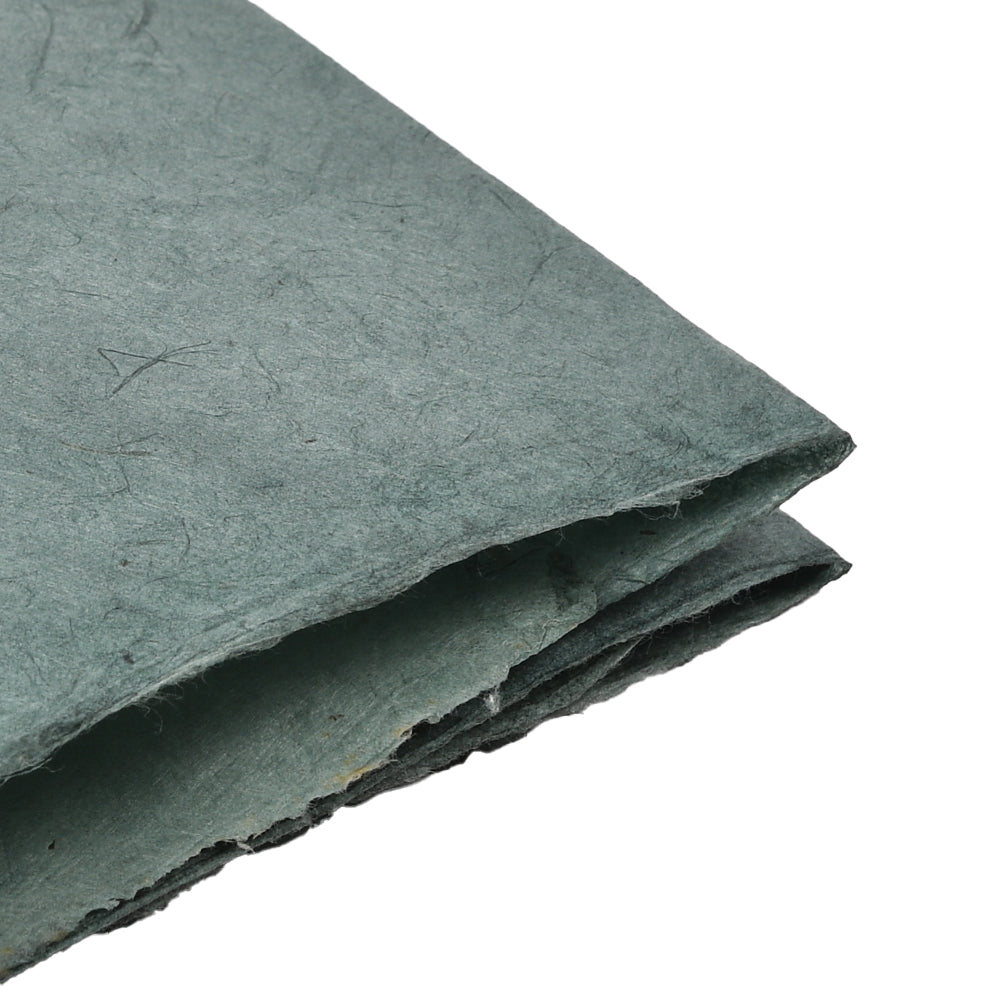 Nahaufnahme von gefaltetem, grünem, handgeschöpftem Nepalpapier in Antikgrün, das seine raue, faserige Struktur vor einem weißen Hintergrund zur Geltung bringt. Dieses nachhaltige Papier unterstreicht sowohl die einzigartige Handwerkskunst als auch seine Umweltfreundlichkeit.