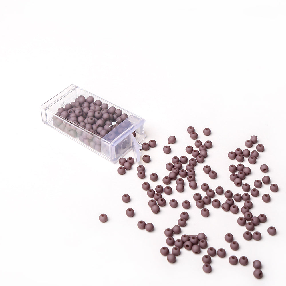 Kleine braune Acrylperlen von Alexandra Renke schwappen aus einer durchsichtigen Plastikbox auf eine weiße Fläche, bereit, Bastelprojekte und Schmuckherstellung zu inspirieren.