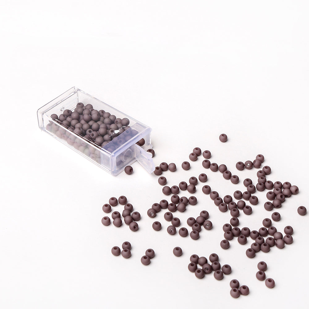 Kleine braune Acrylperlen von Alexandra Renke schwappen aus einem durchsichtigen Kunststoffbehälter auf eine weiße Fläche - ideal für Bastelprojekte oder zum Basteln einzigartiger Schmuckstücke.