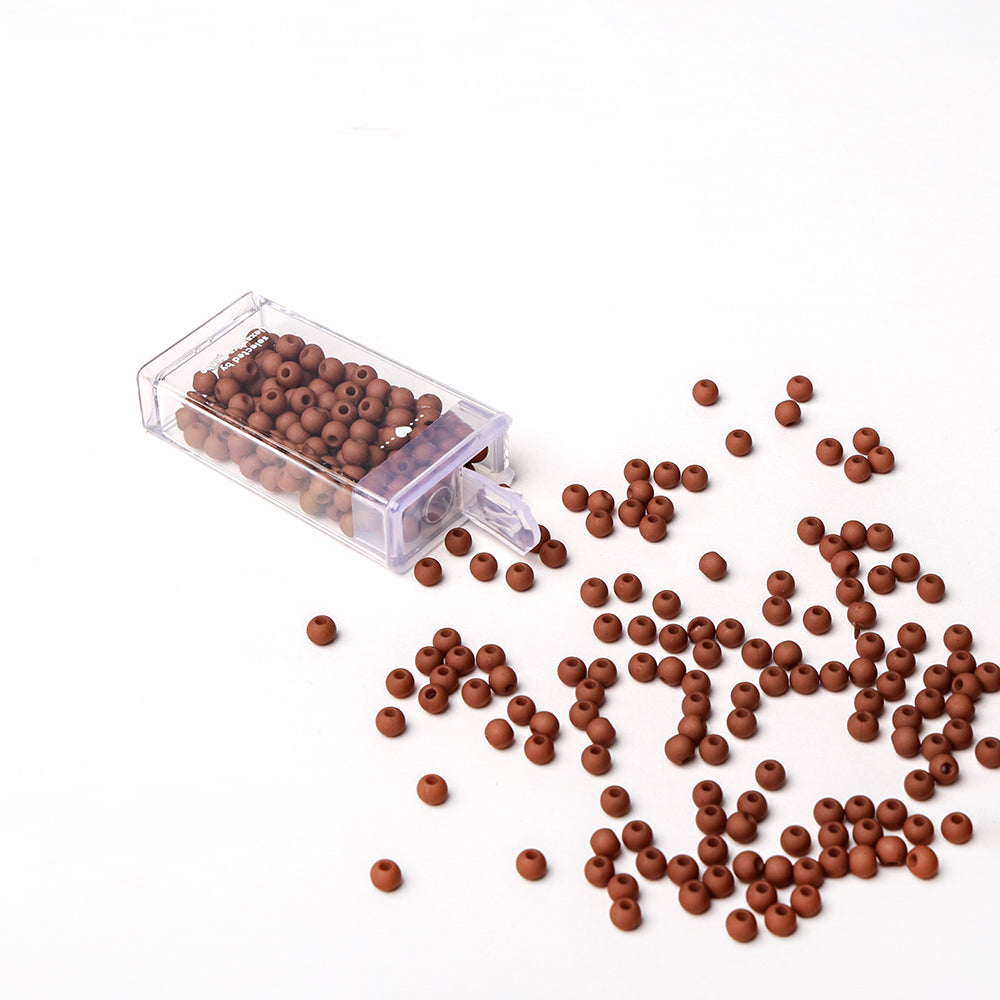 Kleine braune Acrylperlen von Alexandra Renke schwappen aus einem durchsichtigen Kunststoffbehälter auf eine weiße Fläche - ideal für Bastelprojekte oder zum Basteln von einzigartigen Schmuckstücken.