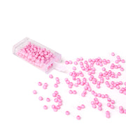 Kleine rosafarbene Acrylperlen von Alexandra Renke werden aus einem durchsichtigen Kunststoffbehälter auf weißem Hintergrund verstreut, ideal für Schmuck basteln und kreative Bastelprojekte.