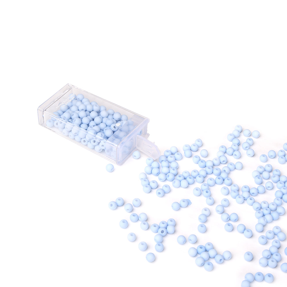 Kleine blaue Acrylperlen von Alexandra Renke, die aus einem klaren Gefäß auf eine weiße Fläche tropfen, ideal für Schmuck basteln und kreative Bastelprojekte.