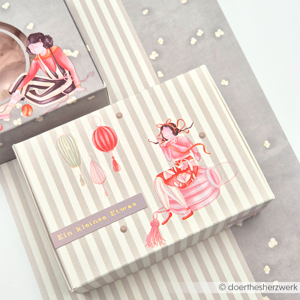 Ein gestreifter Geschenkkarton mit skurrilen Frauen- und Ballonillustrationen, hergestellt mit Alexandra Renke Rub-On Figuren, liegt inmitten kleiner weißer Konfettis - perfekt für Ihr Bastelprojekte Motiv.