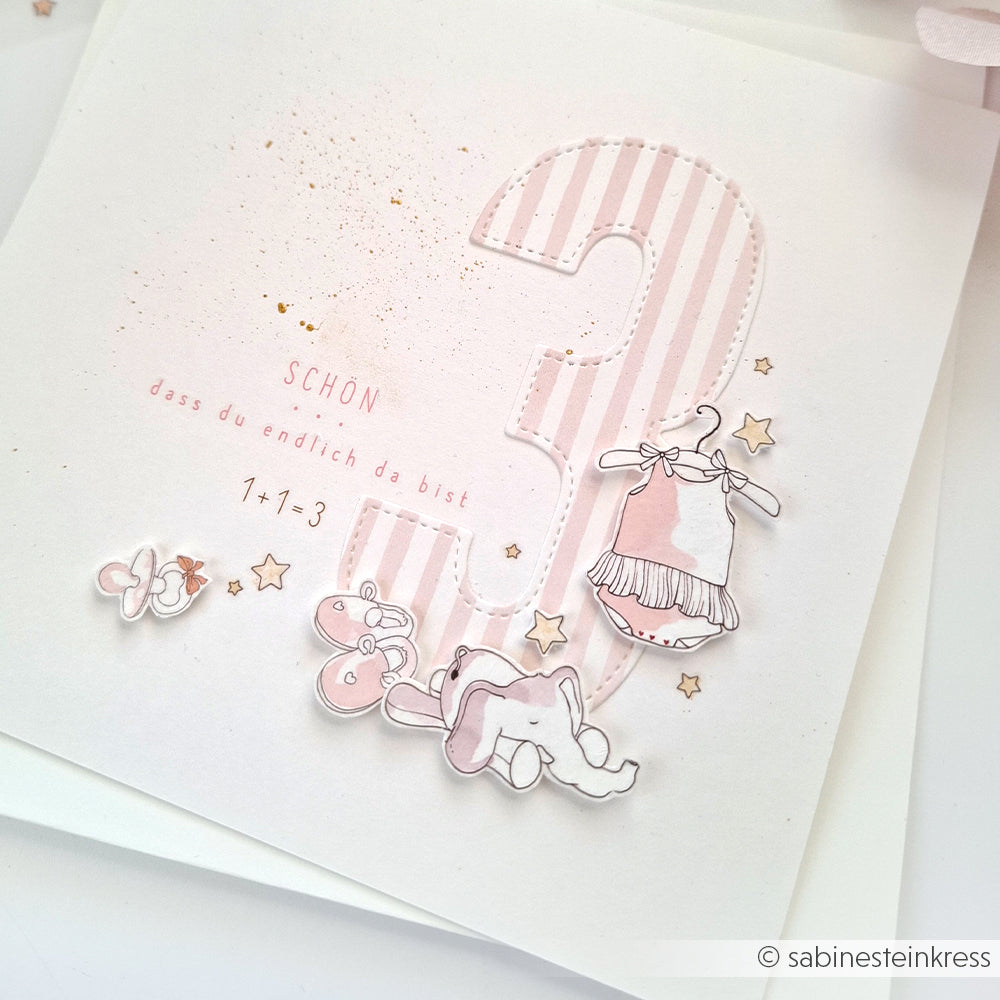 Die rosa-weiße Karte von Alexandra Renke zeigt eine große gestreifte Zahl 3 mit Motiven von Babykleidung, Schuhen und Spielzeug sowie deutschem Text - ideal für Bastelprojekte oder zum Verzieren mit Transferfolien-Basteln.