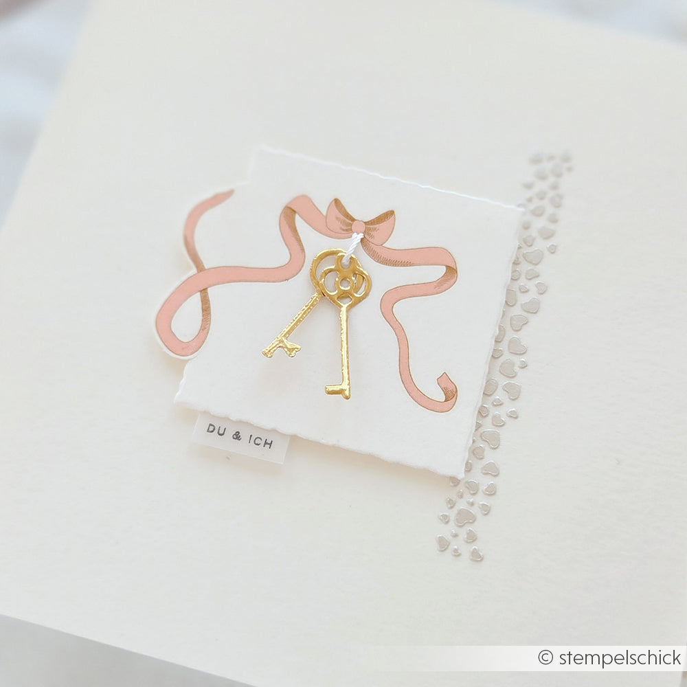 Ein goldener Schlüssel und ein herzförmiger Schlüssel auf einer Karte mit rosafarbener Schleife und DU & ICH-Etikett von Alexandra Renke - ideal für Bastelprojekte oder zur Verwendung mit Rubbeltransferfolie.