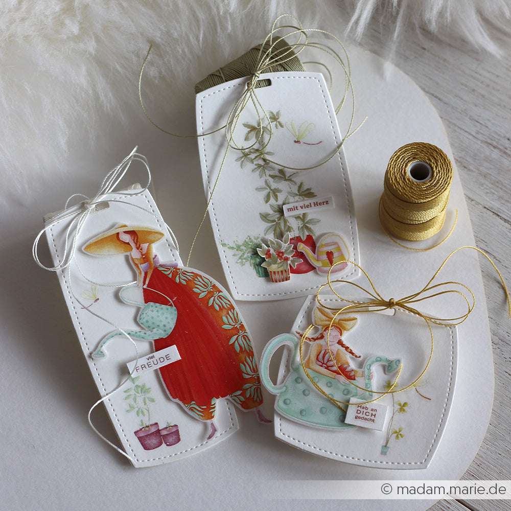 Drei dekorative Geschenkanhänger von Alexandra Renke mit Motiven wie Frauen, Teeservice und Pflanzen, gebunden mit weiß-goldenem Zwirn - ideal für kreative Geschenke mit Transferfolien Basteln.