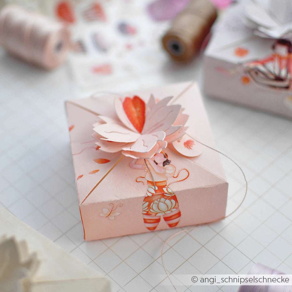 Eine pastellrosa Papier-Geschenkbox von Alexandra Renke mit Blumen- und Buchstabenmotiven, verziert mit Papierblumen und Faden - ideal für Bastelprojekte Motive oder zur Aufwertung von Projekten mit Rub-On Figurenwelt.
