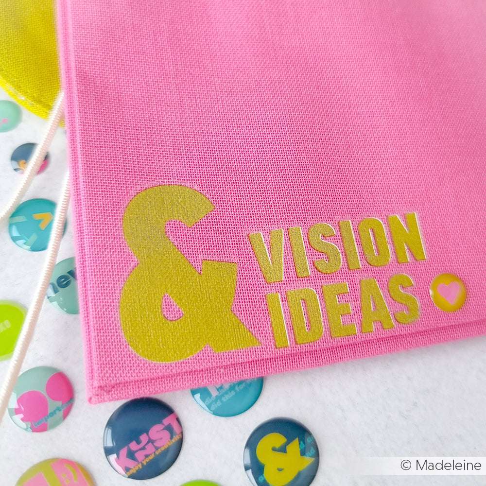 Der rosafarbene Stoff von Alexandra Renke mit der Aufschrift & VISION IDEAS" in gelben Lettern sitzt neben bunten runden Knöpfen - ideal für Bastelprojekte oder zum Hinzufügen von Motiven aus Rub-On Transferfolien.